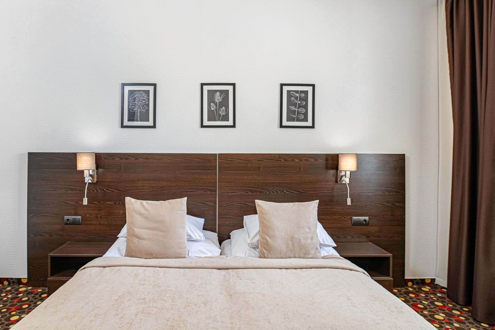 Een tweepersoonsbed met een houten hoofdbord, twee nachtkastjes met lampen en drie ingelijste botanische prints aan de muur.