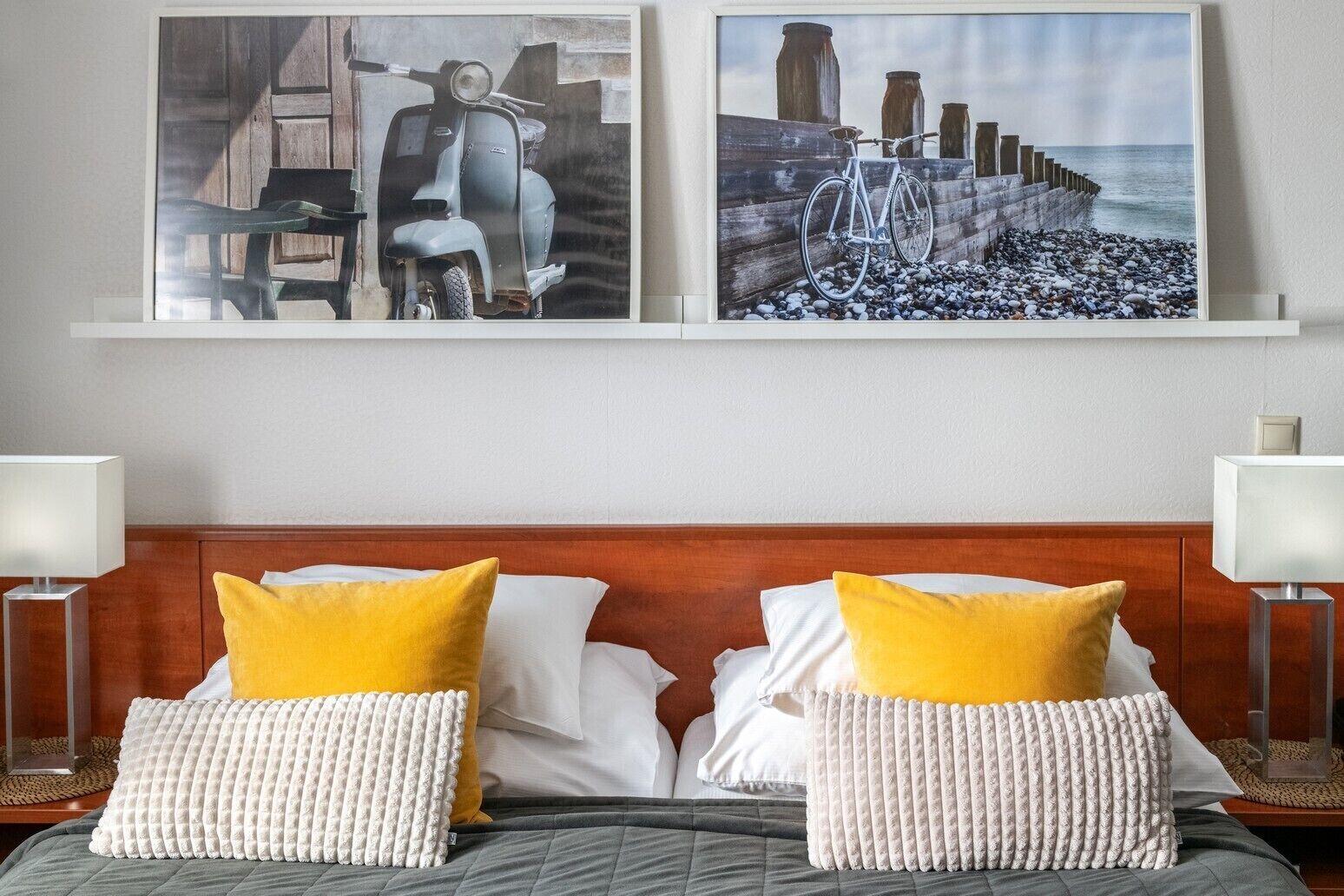 A hotel room with a wooden headboard, two beds with white and yellow pillows, and two framed artworks above the beds.