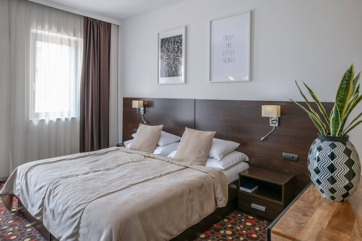 A hotel room with a large bed, two bedside tables, a potted plant, and artwork on the walls.