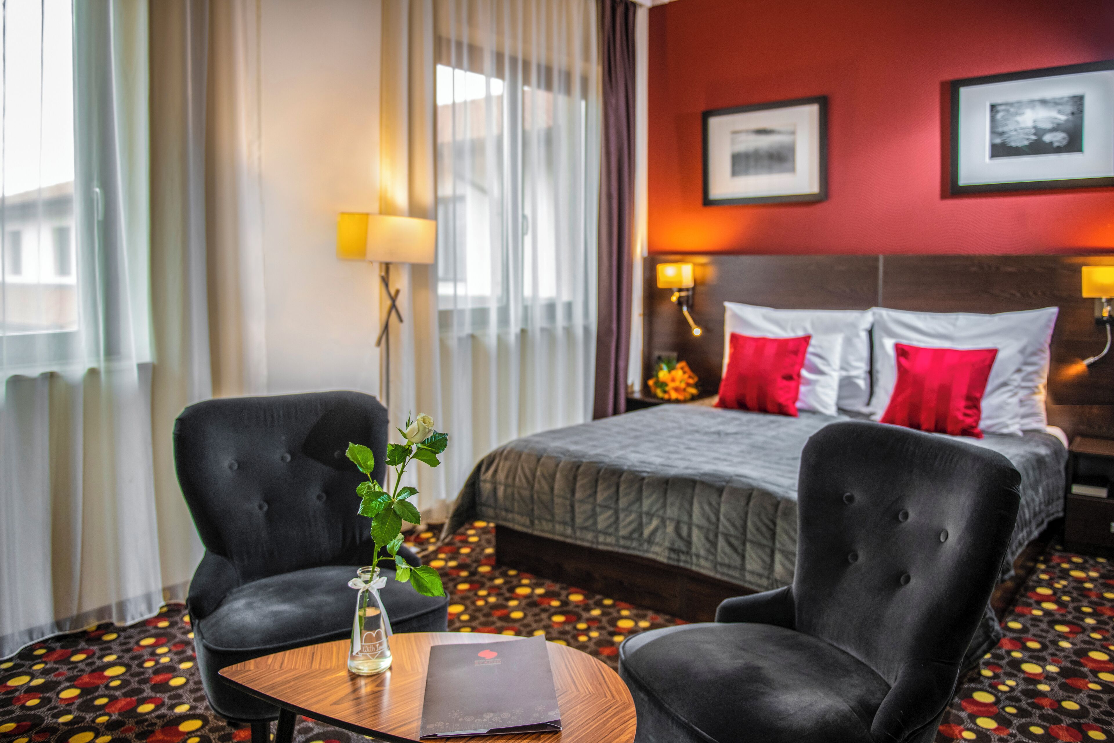 Habitación de hotel con cama, dos sillones, una mesita con un jarrón y un libro, y una alfombra estampada.