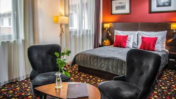 Habitación de hotel con cama, dos sillones, una mesita con un jarrón y un libro, y una alfombra estampada.