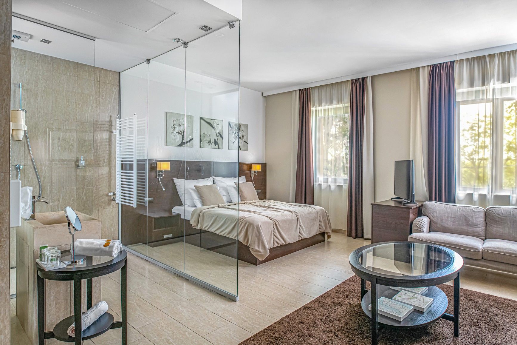 Una habitación de hotel moderna con cabina de ducha de vidrio, cama con cabecero, sofá, mesa de centro y una obra de arte en la pared.