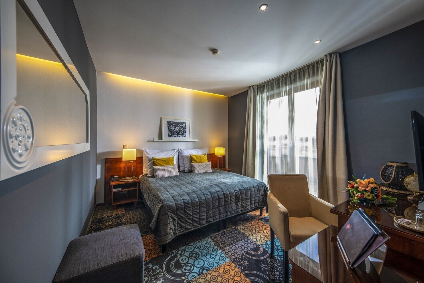 A hotel room with a bed, bedside tables, a chair, a desk, and a large window with curtains.