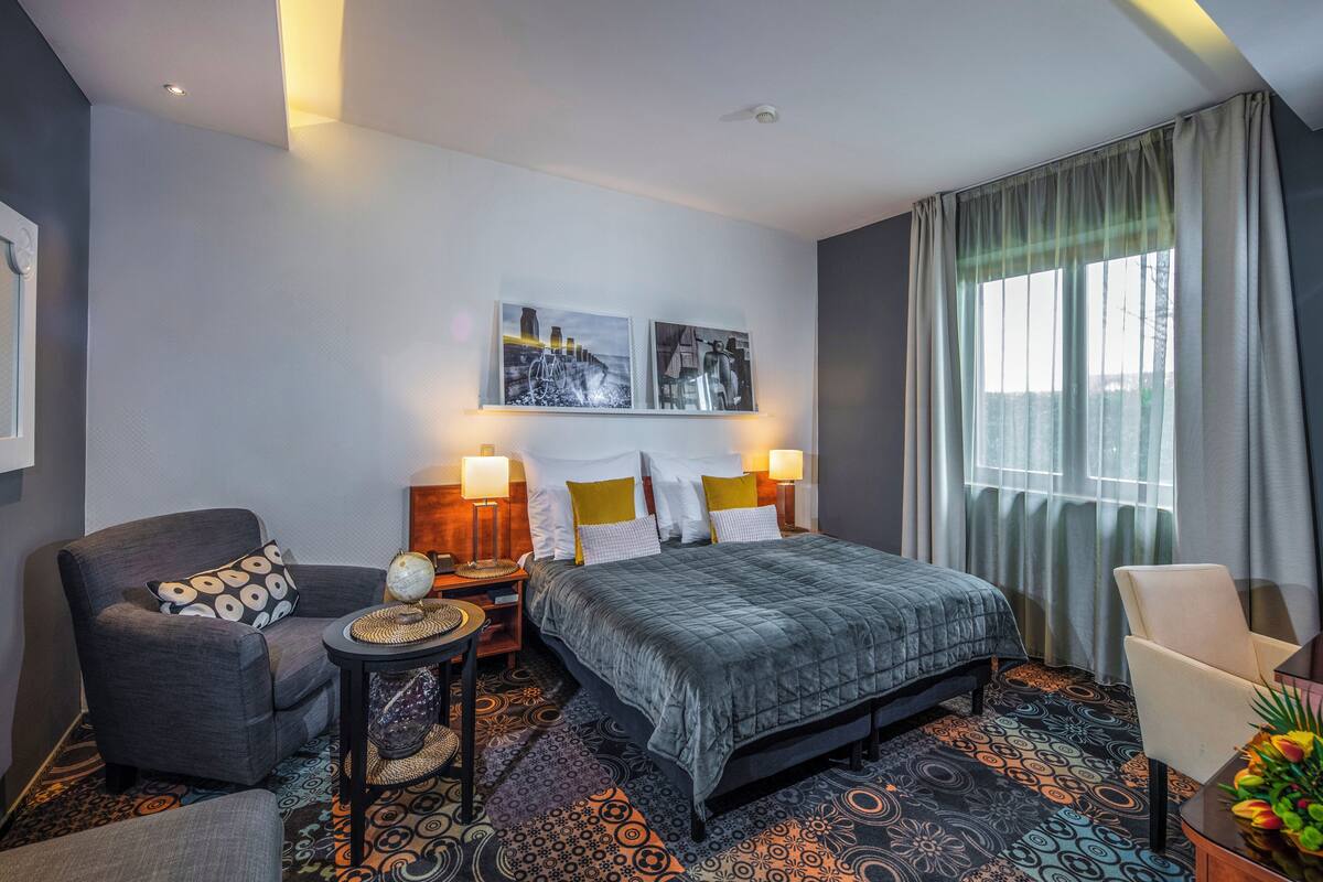 A hotel room with a bed, two chairs, a nightstand, a lamp, and a window with curtains.