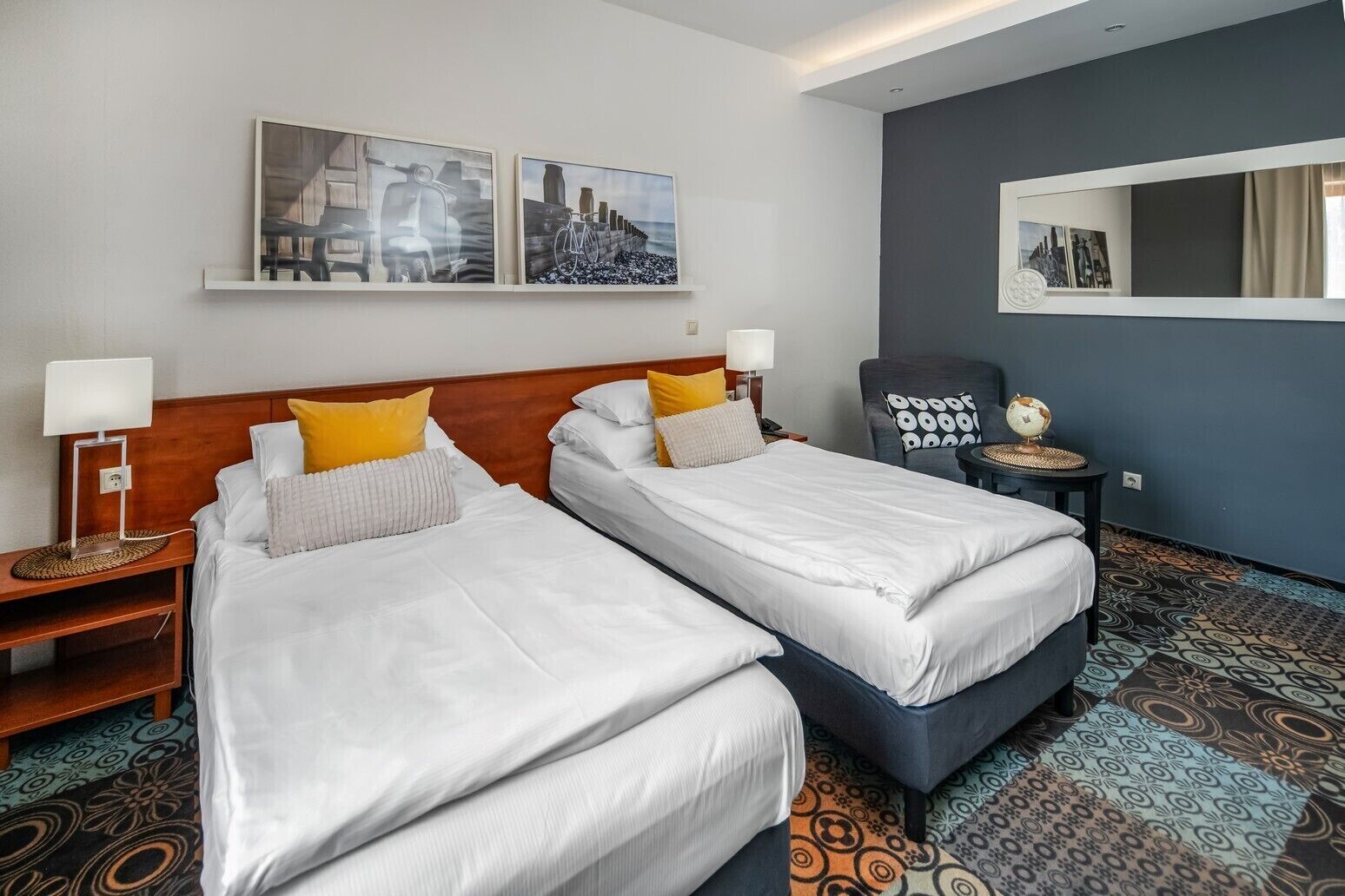 Une chambre d’hôtel avec deux lits, une tête de lit en bois, un miroir, une chaise, une petite table sur laquelle se trouve un globe terrestre et une moquette à motifs.