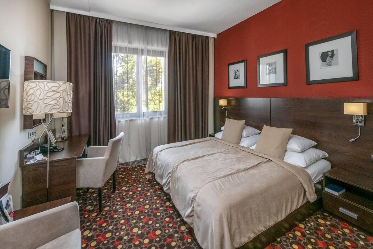 A hotel room with a bed, a desk, a chair, a window with curtains, and artwork on the walls.