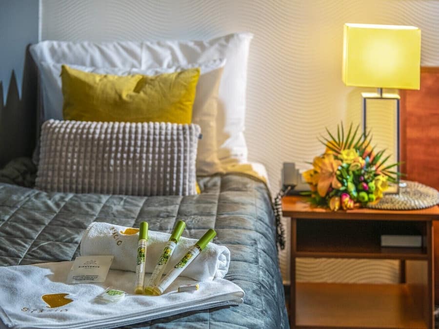 A neatly made bed with a quilted cover, a white pillow, a green pillow, and a decorative pillow with a checkered pattern. There is a bedside table with a lamp, a floral arrangement, and a folded towel with a card on it.