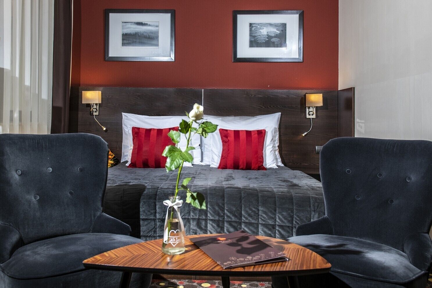 Una habitación de hotel con una cama grande, dos lámparas de noche, un jarrón con una flor y dos sillones.