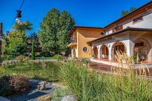 A house with a pond, surrounded by greenery and a clear blue sky. - Budapest Airport Hotel Stáció Wellness & Conference (Vecsés)