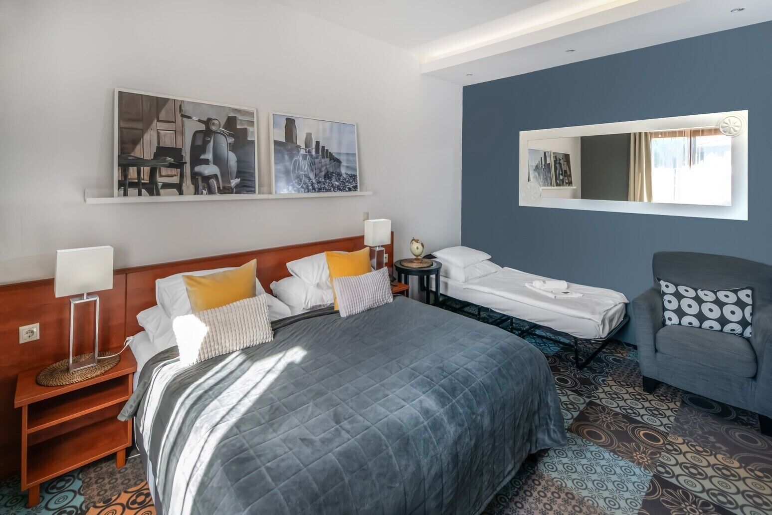 A hotel room with a large bed, a smaller bed, a desk, a chair, and artwork on the walls.