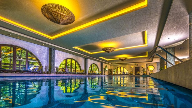 A modern indoor swimming pool with clear water, yellow lighting accents, and arched windows.