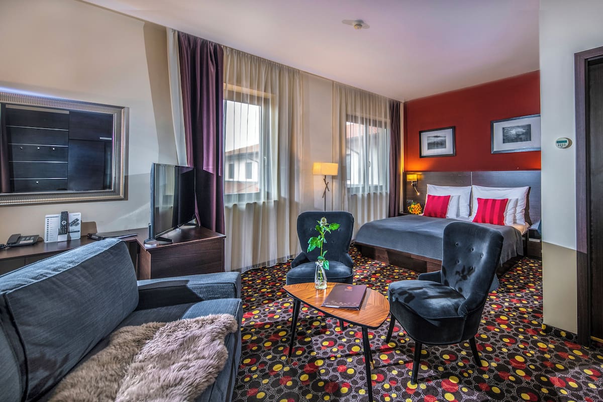 A hotel room with a bed, a sofa, a chair, a desk with a lamp, a television, and a patterned carpet.