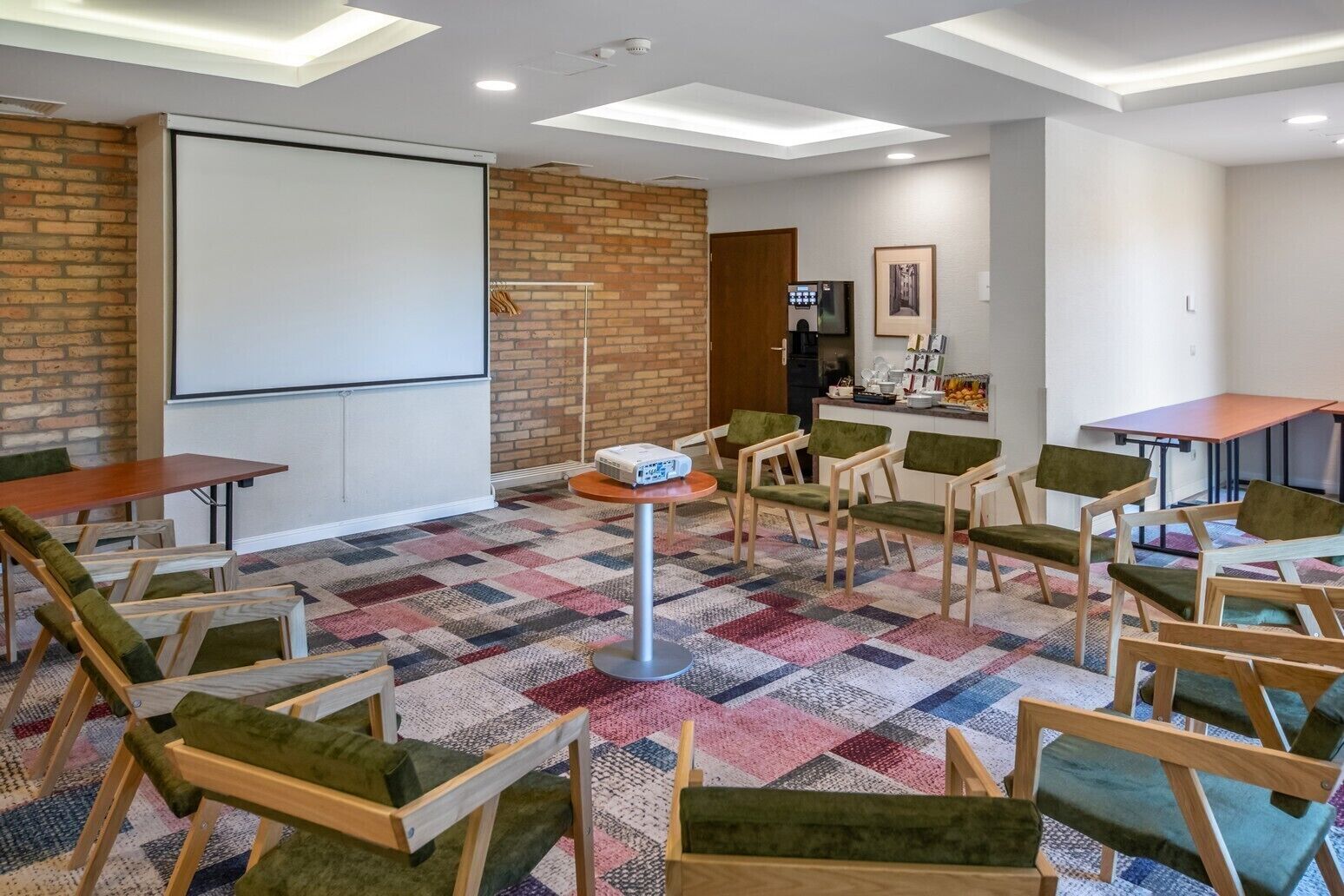 Una sala de conferencias con pantalla de proyector, pared de ladrillo, kitchenette y alfombra estampada.