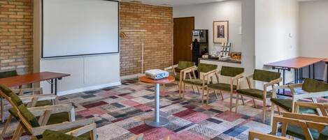 A conference room with a projector screen, a brick wall, a kitchenette, and a patterned carpet.
