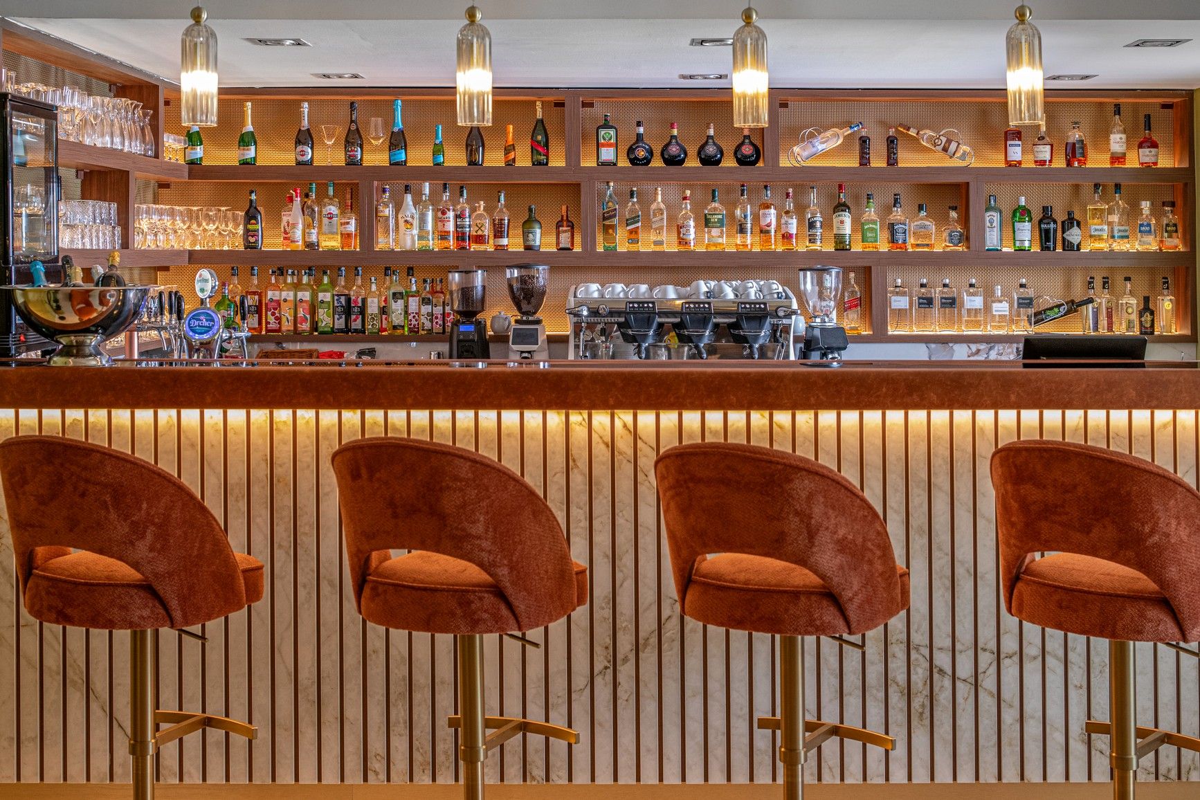Uma área de bar com várias prateleiras de garrafas de bebidas, um balcão e três banquetas estofadas.