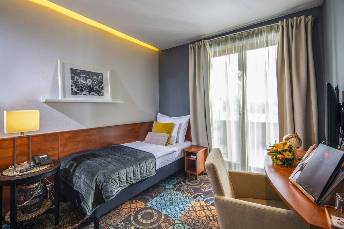 A hotel room with a bed, bedside table, lamp, chair, desk with a computer, and a window with curtains.