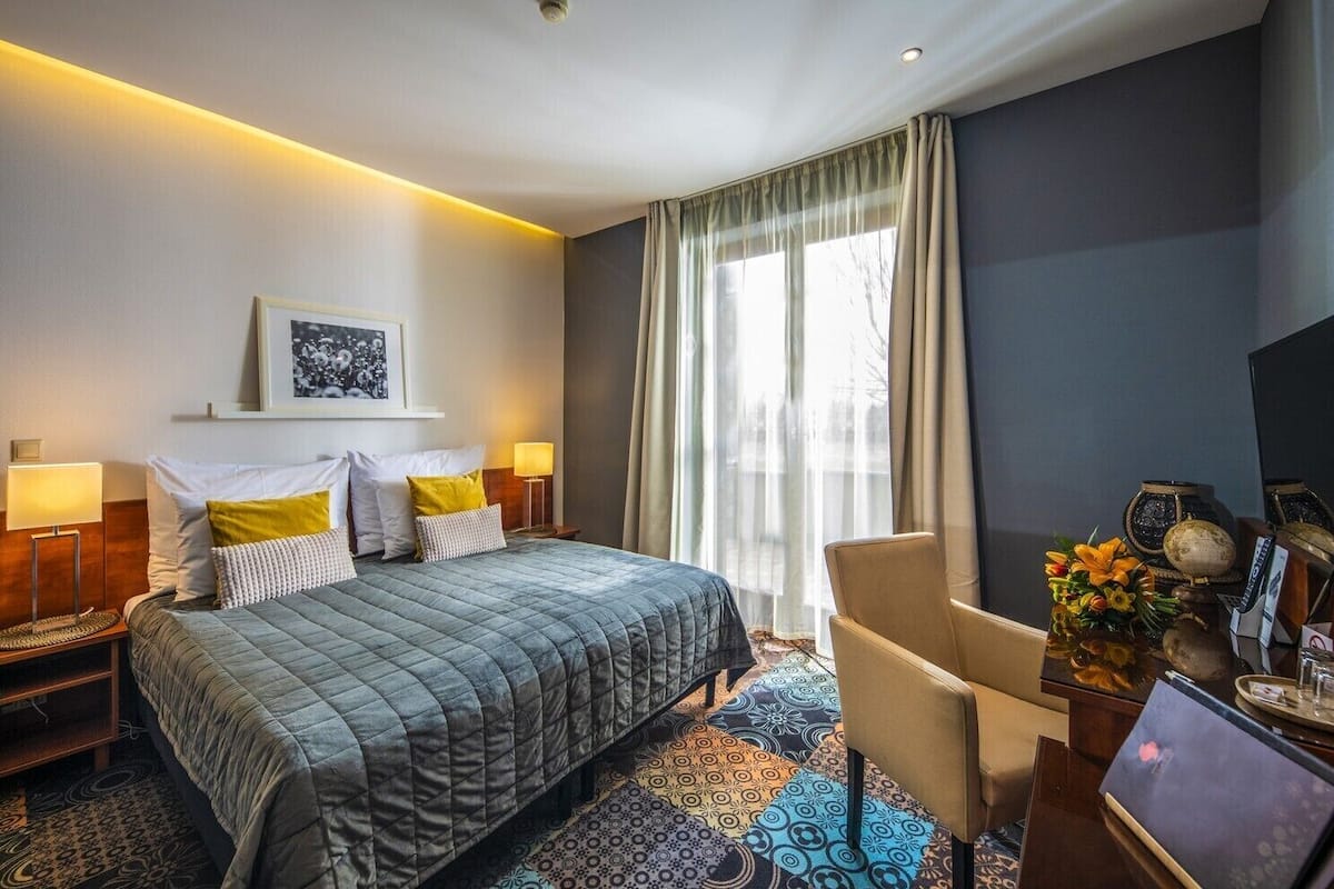 A hotel room with a bed, two bedside lamps, a desk with a vase of flowers, and a large window with curtains.
