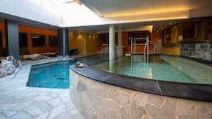 Una piscina interior con paredes y suelo de piedra, rodeada por una terraza de piedra y otra de madera con asientos.