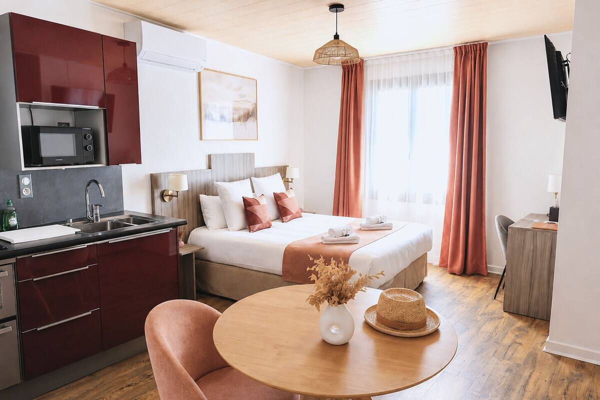 A modern hotel room with a kitchen area, a bed with a wooden headboard, a round wooden table, and a single chair.