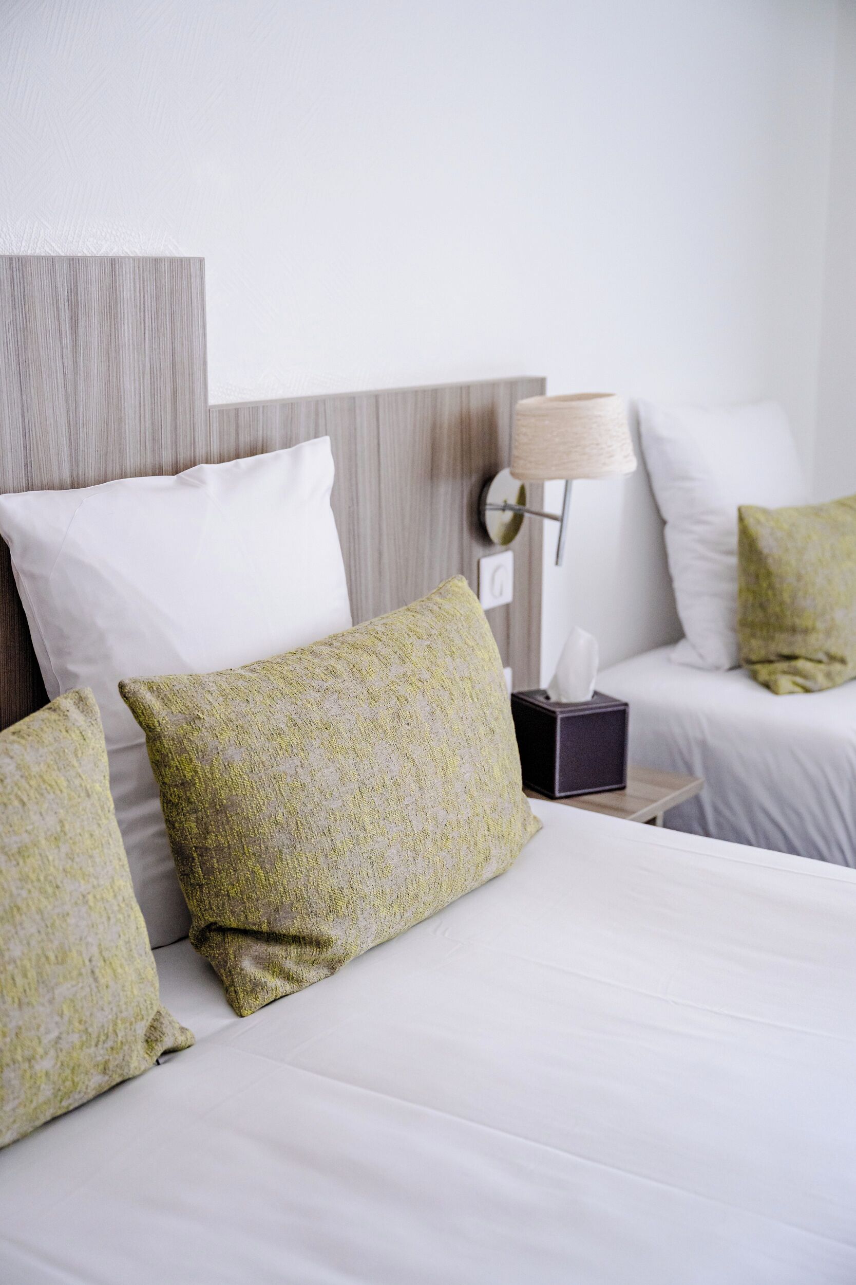 Una cama bien hecha con sábanas blancas y almohadas estampadas en verde, una lámpara de noche y una caja de pañuelos en la mesita de noche.