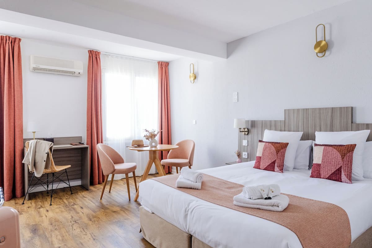 A hotel room with a bed, a desk, a chair, a table, and an air conditioning unit.