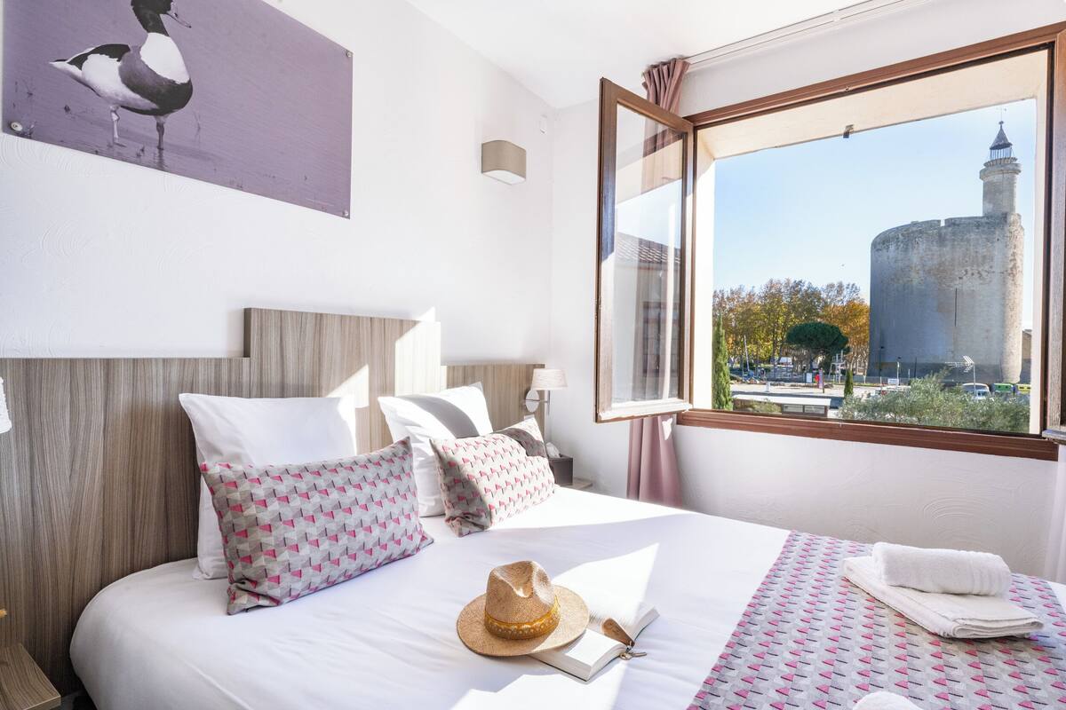 A hotel room with a bed, pillows, a hat, a book, and a view of a tower through a window.