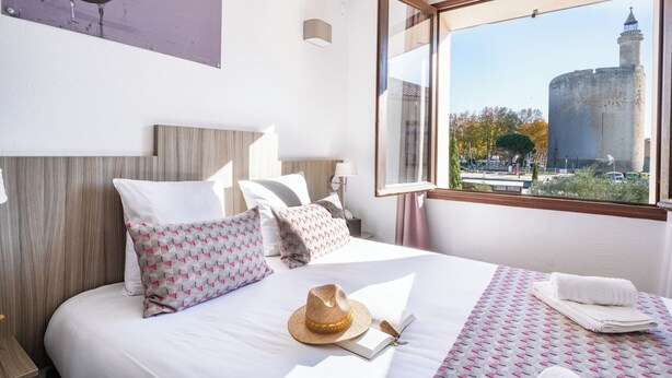 Habitación de hotel con cama, almohadas, un sombrero, un libro y vista a una torre a través de una ventana.