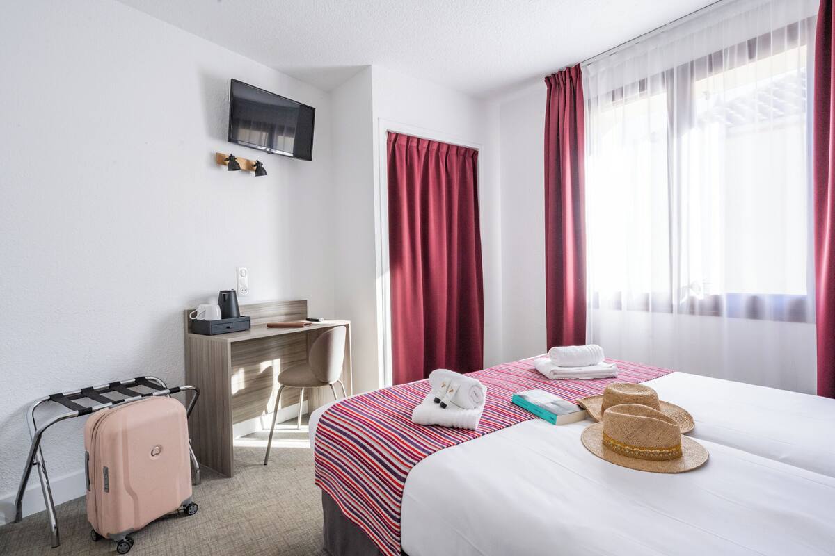 A hotel room with a bed, a desk, a TV, a suitcase, and a window with curtains.