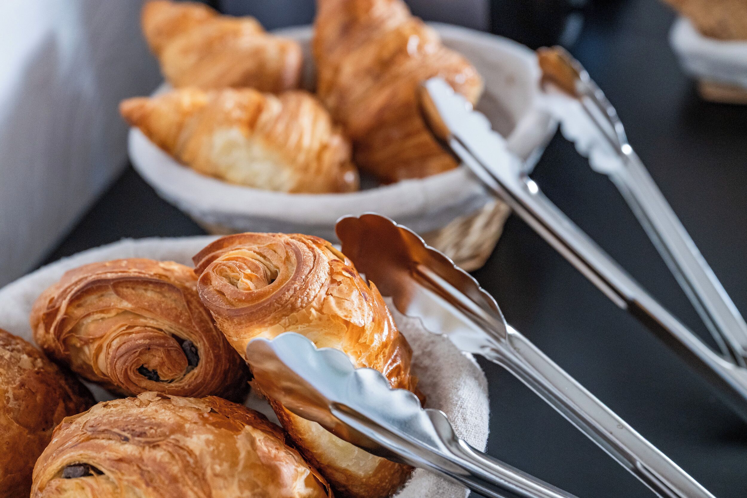 A close-up of a basket filled with croissants and a pair of tongs.