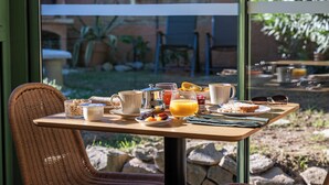 Una mesa de desayuno con diversos alimentos y bebidas, una silla de mimbre y vistas al exterior a través de un ventanal.