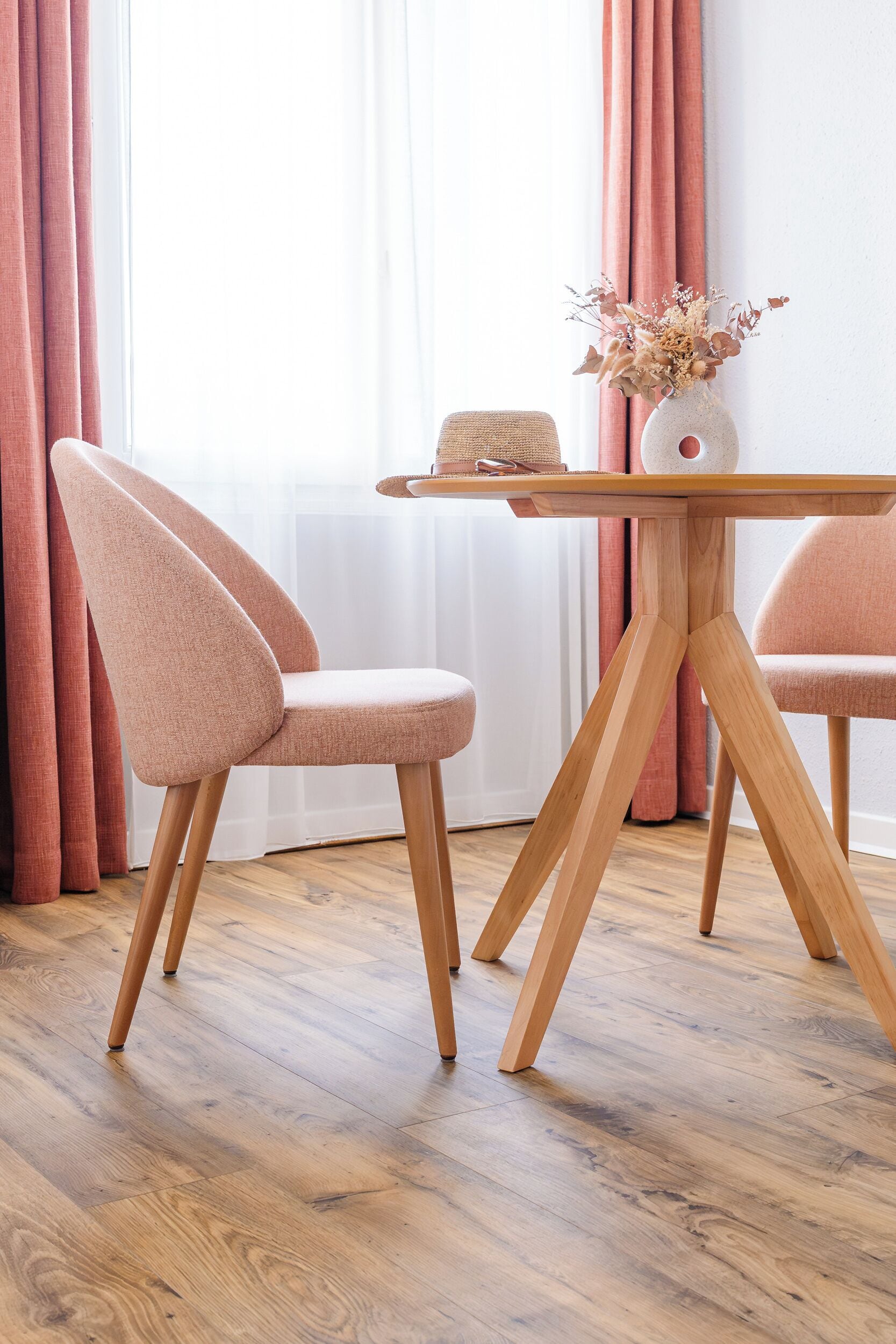 Ein Holztisch mit einer Vase getrockneter Blumen, zwei passende Stühle und ein Fenster mit Vorhängen.