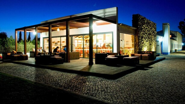 A modern building with large glass windows, outdoor seating, and a cobblestone ground. - Quinta do Mel (Albufeira)