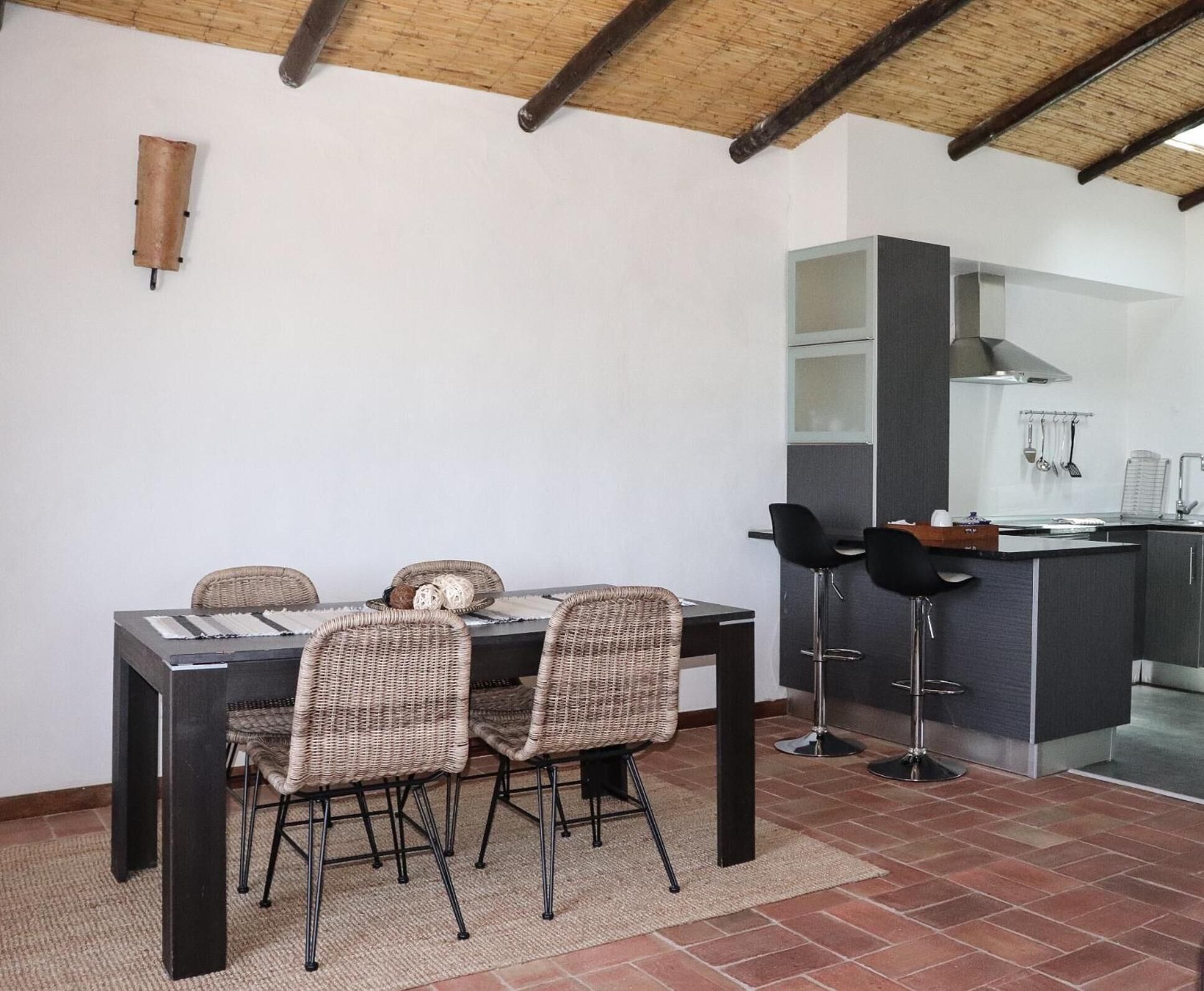 Un comedor con una mesa y cuatro sillas de mimbre, una cocina con barra, y piso de baldosas.