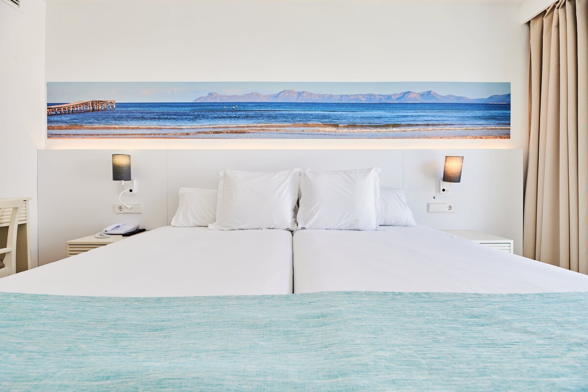Una cama grande con ropa de cama blanca, dos lámparas de noche y un cuadro con una escena costera.