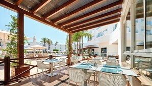 An outdoor patio area with white wicker chairs and tables, a pool in the background, and a modern building facade. - Aluasun Continental Park Hotel & Apartments (Muro)