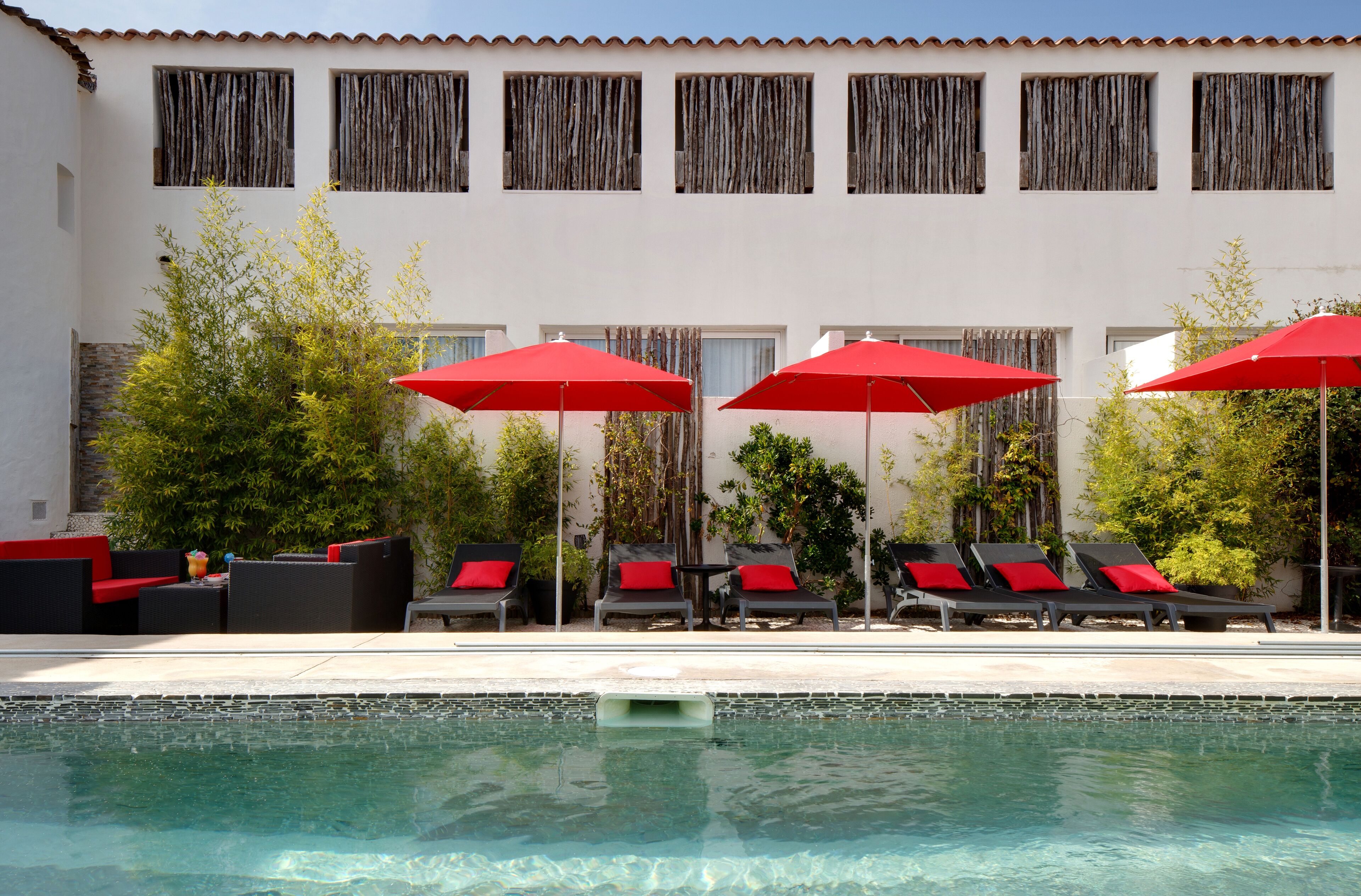 A poolside area with lounge chairs and red umbrellas, a tiled pool, and a building with multiple windows and a tiled roof.