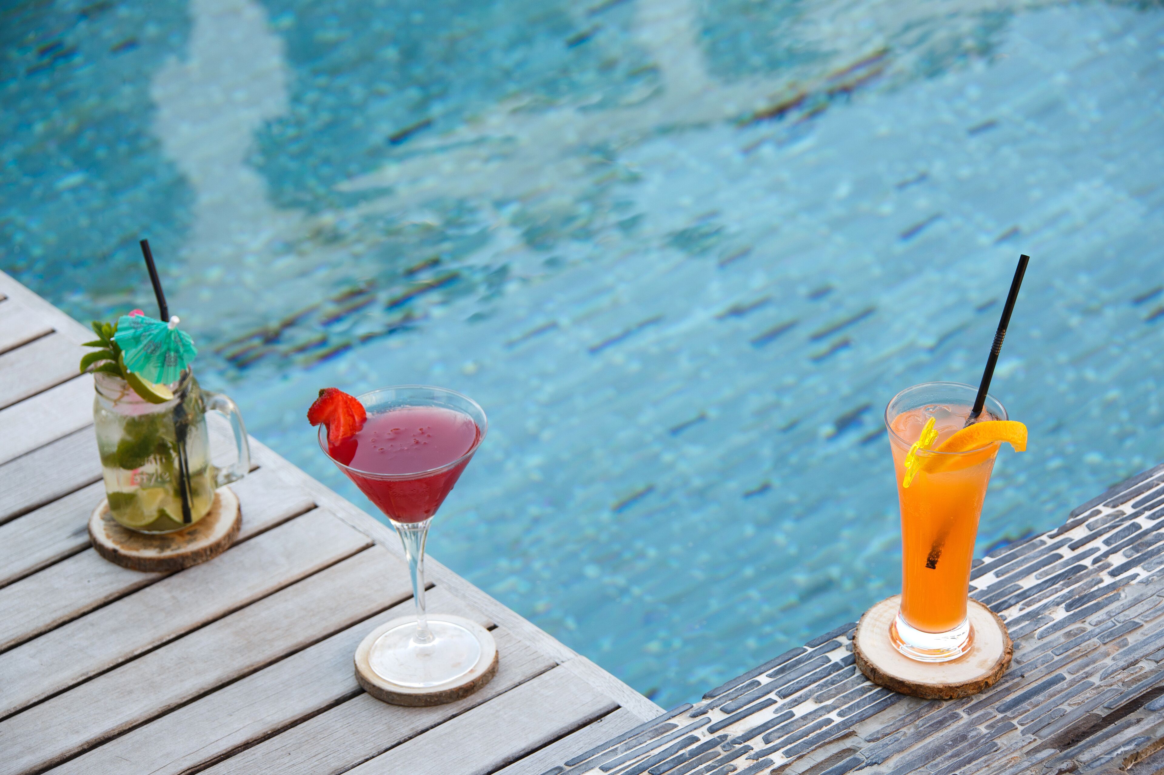 Una terraza de madera con tres bebidas sobre posavasos junto a una piscina.