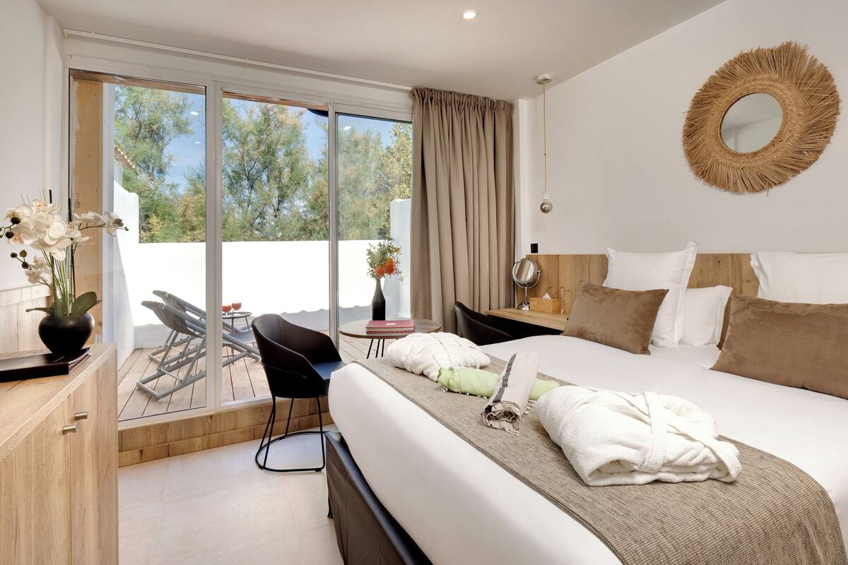 A modern hotel room with a large bed, a wooden headboard, a round mirror, and a view of the outdoors through a sliding glass door.