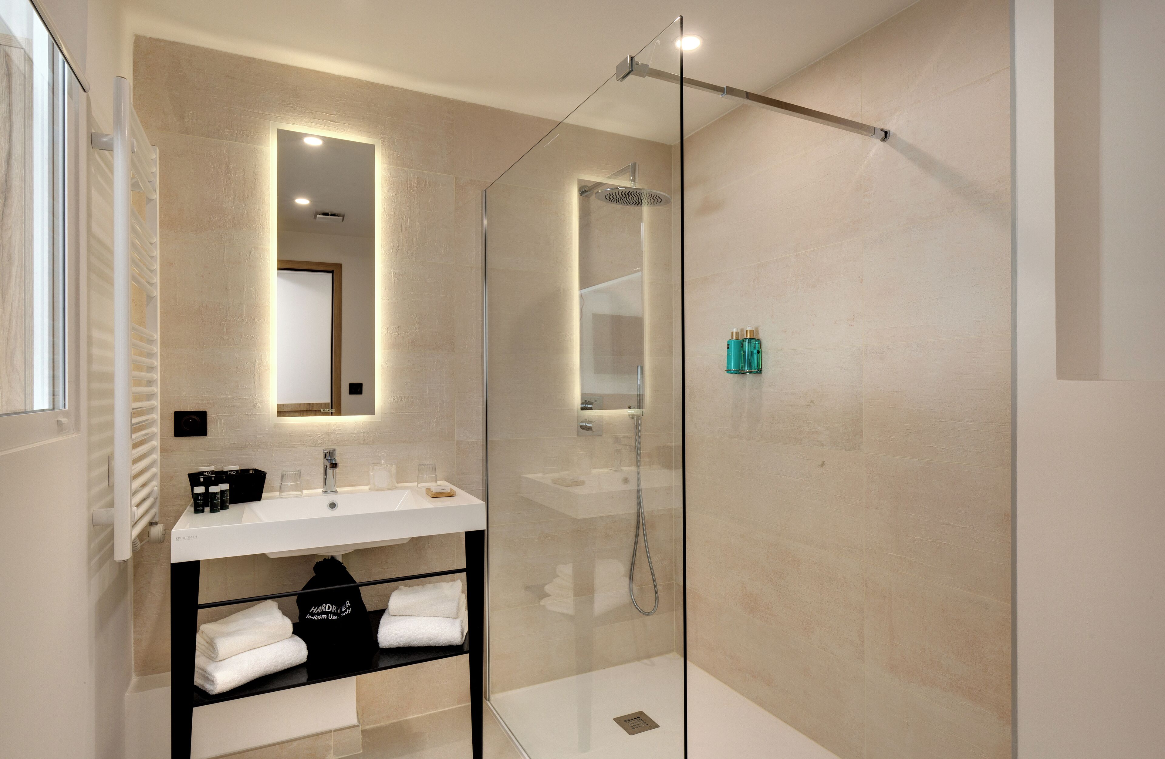 Un baño moderno con ducha de vidrio, un lavamanos blanco con espejo y un estante con toallas y artículos de aseo.