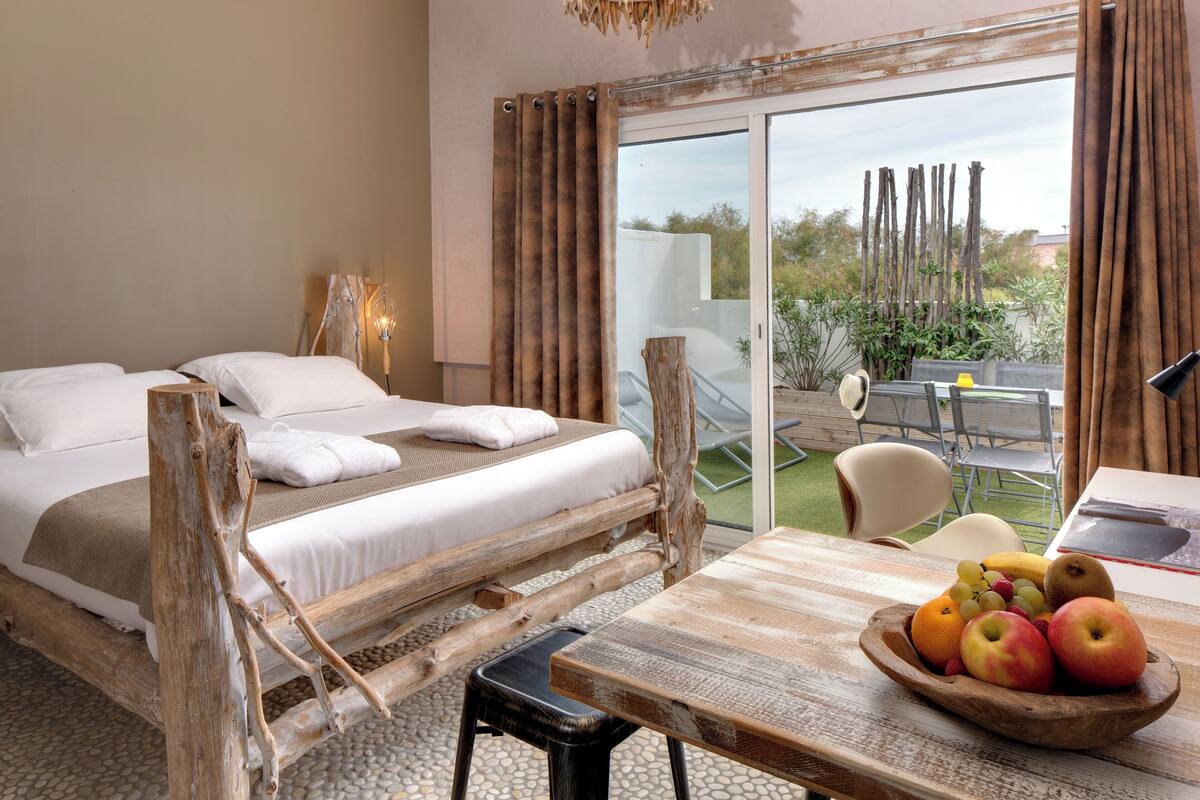 A bedroom with a bed, a wooden table with a fruit bowl, and a view of the outdoors.