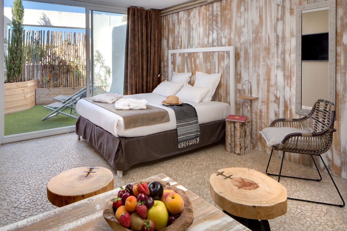 A hotel room with a bed, a dining table with fruit, a wicker chair, and a view of the outdoors.