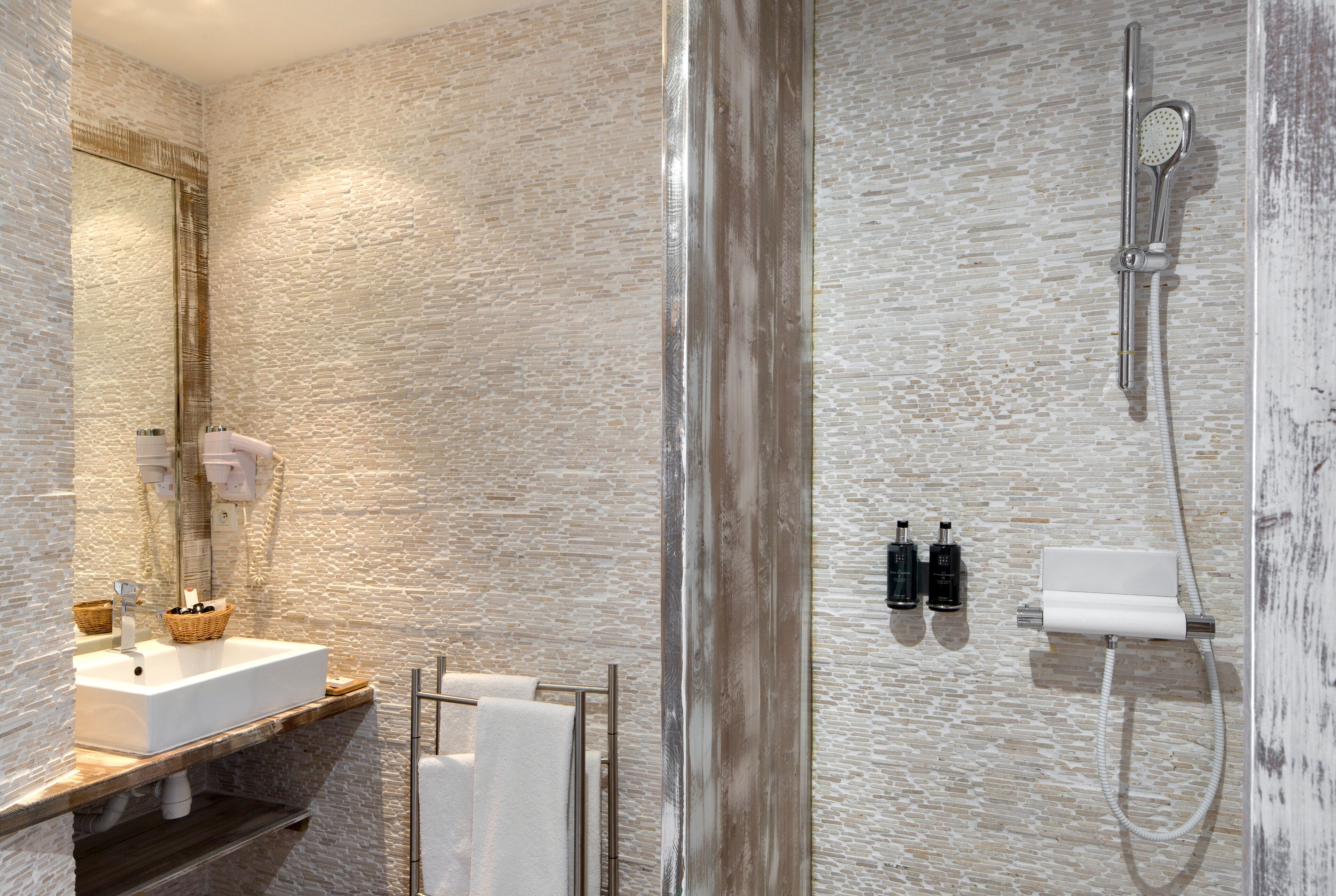 A modern bathroom with a textured stone wall, a wooden vanity with a sink, a glass shower enclosure, and a towel rack.