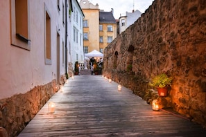 A narrow alleyway with wooden flooring and stone walls, lined with candles and potted plants.