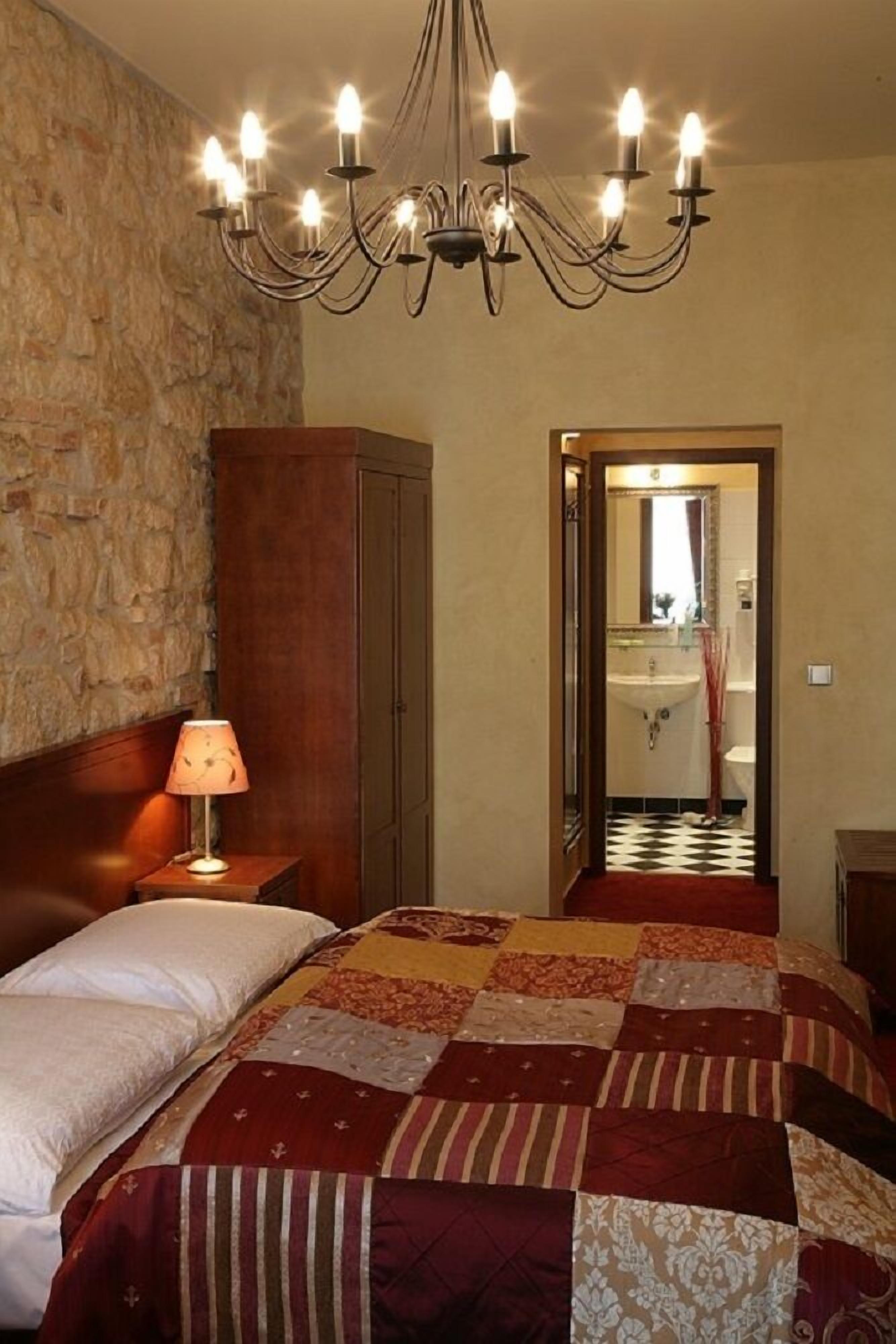 A bedroom with a large bed, a wooden wardrobe, a chandelier, and a bathroom visible through an open door.