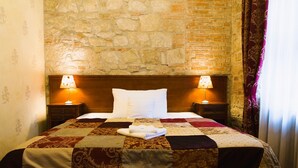 A hotel room with a stone wall, a wooden headboard, a quilted bedspread, two bedside lamps, and a neatly made bed with white pillows. - Hotel Rous (Plzen)