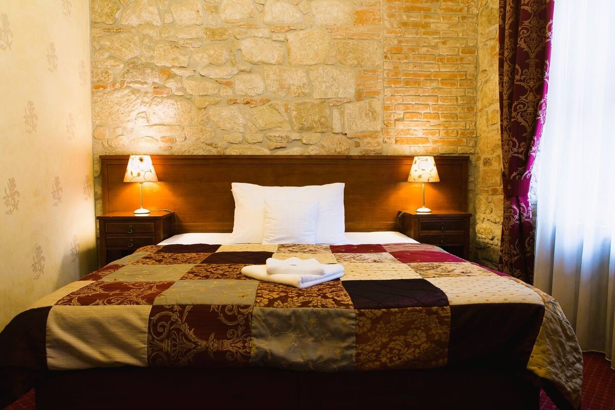 Una habitación de hotel con pared de piedra, cabecero de madera, cubrecama acolchado, dos lámparas de noche y una cama bien hecha con almohadas blancas.