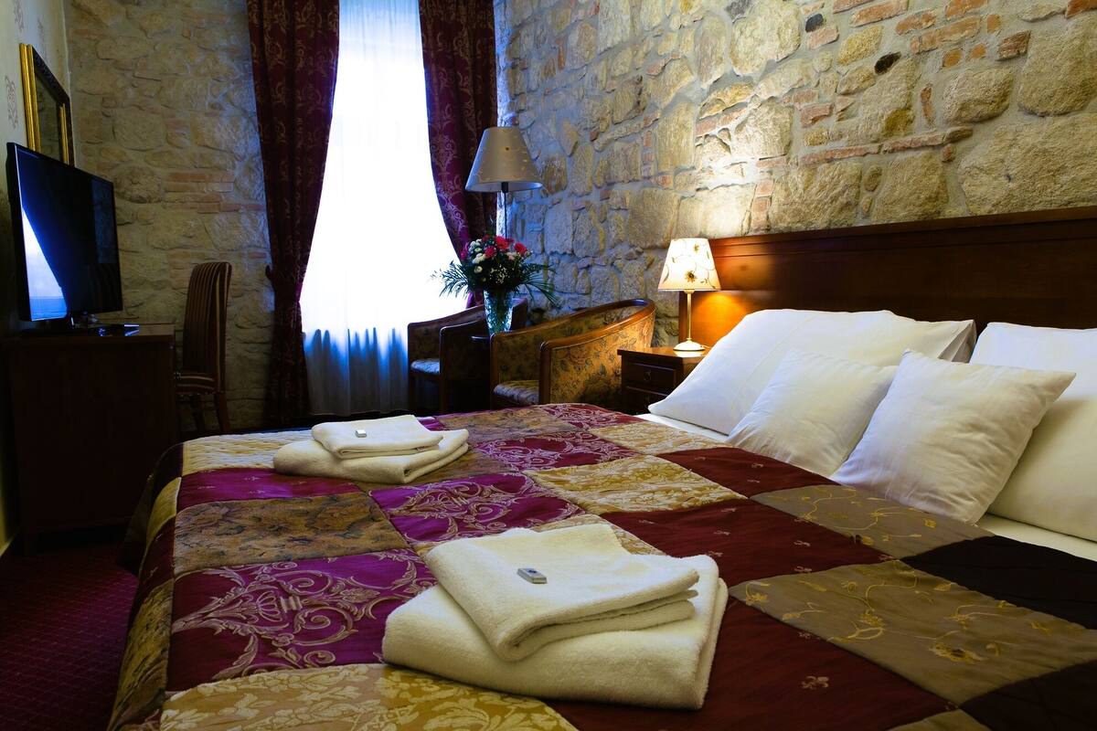 Una habitación de hotel con pared de piedra, una cama con colcha, un escritorio con televisor y una mesita de noche con lámpara.