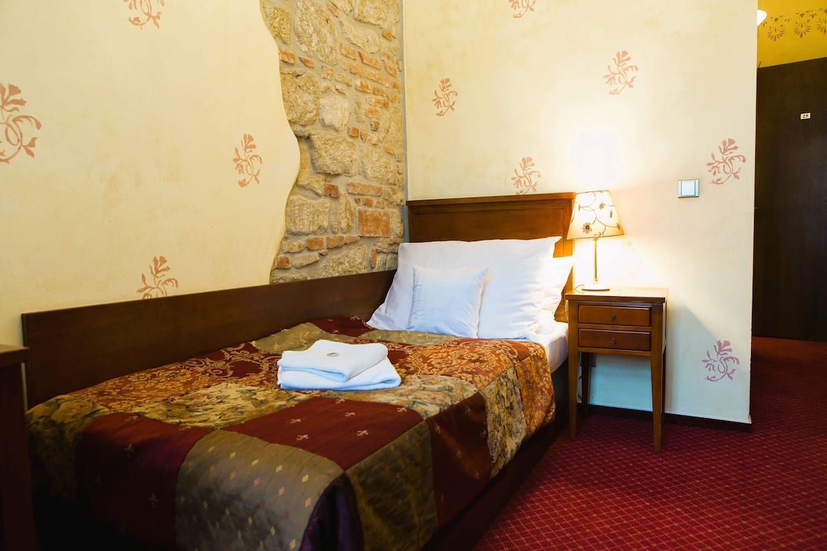 Habitación de hotel con pared de piedra, cama con colcha estampada, almohadas blancas, mesita de noche con lámpara y alfombra roja.