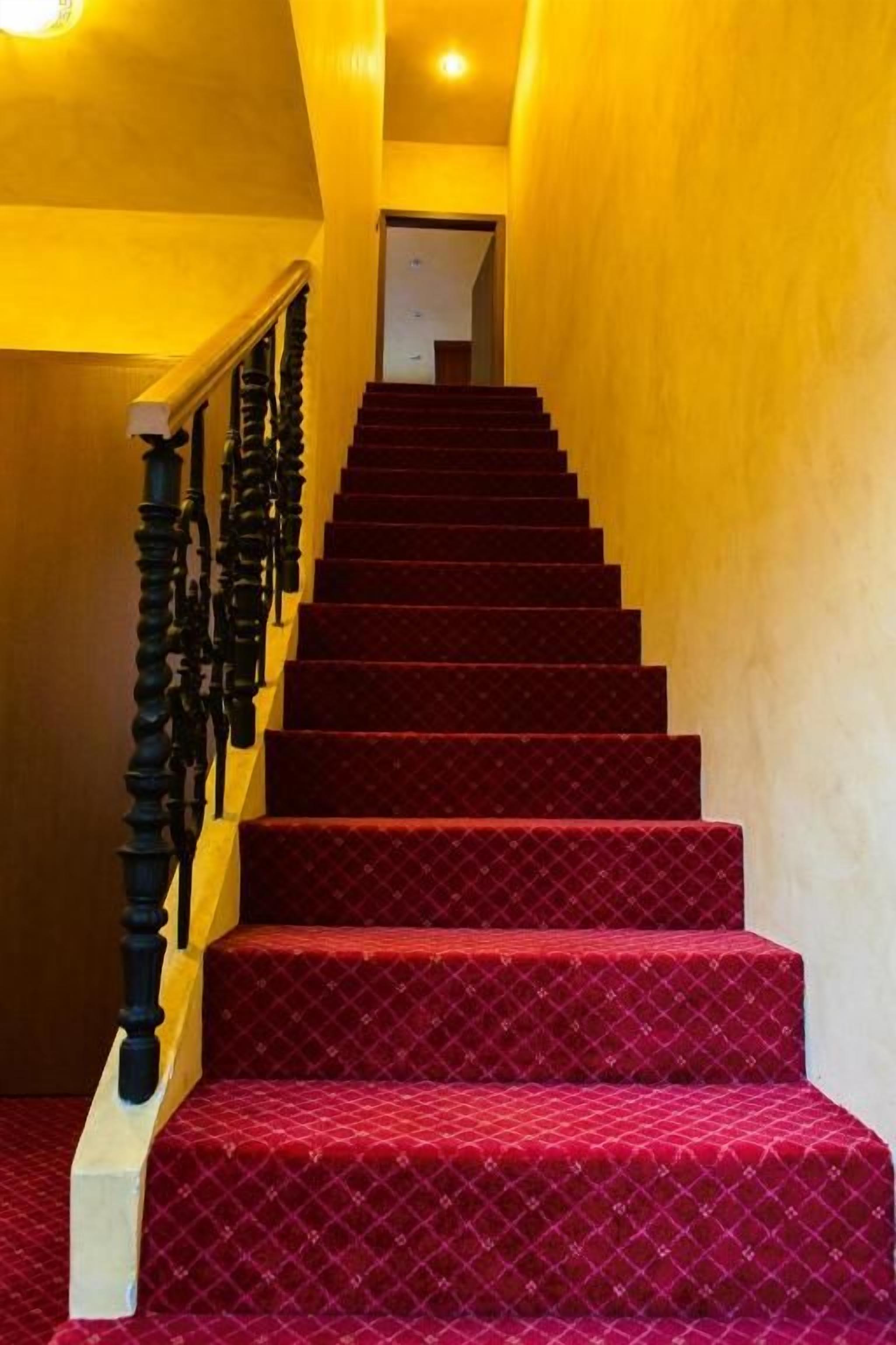 Escadaria com degraus carpetados em vermelho e corrimão preto, levando a uma porta no topo.