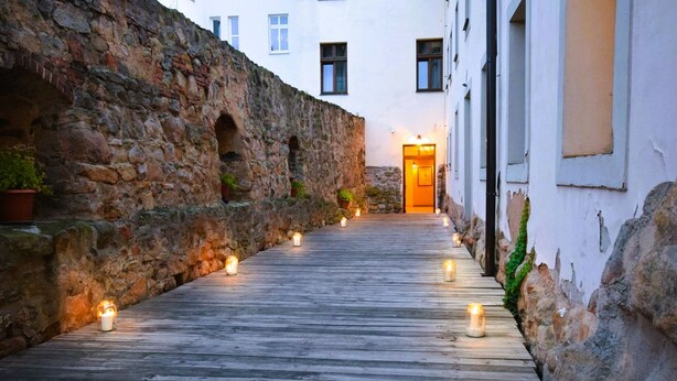 A narrow wooden pathway flanked by stone walls, leading to a building entrance with lit candles. - Hotel Rous (Plzen)